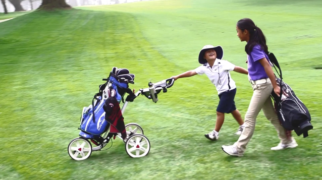 Two golfers, one male and one female, walk a golf course green with each other while rolling a club cart